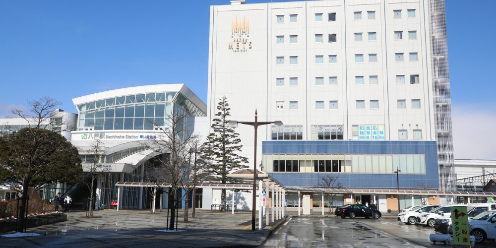 青空の下、左手にガラス張りのJR八戸駅駅舎と右手に高層ホテルが並び、手前にタクシー乗降所や歩道が広がる駅前風景
