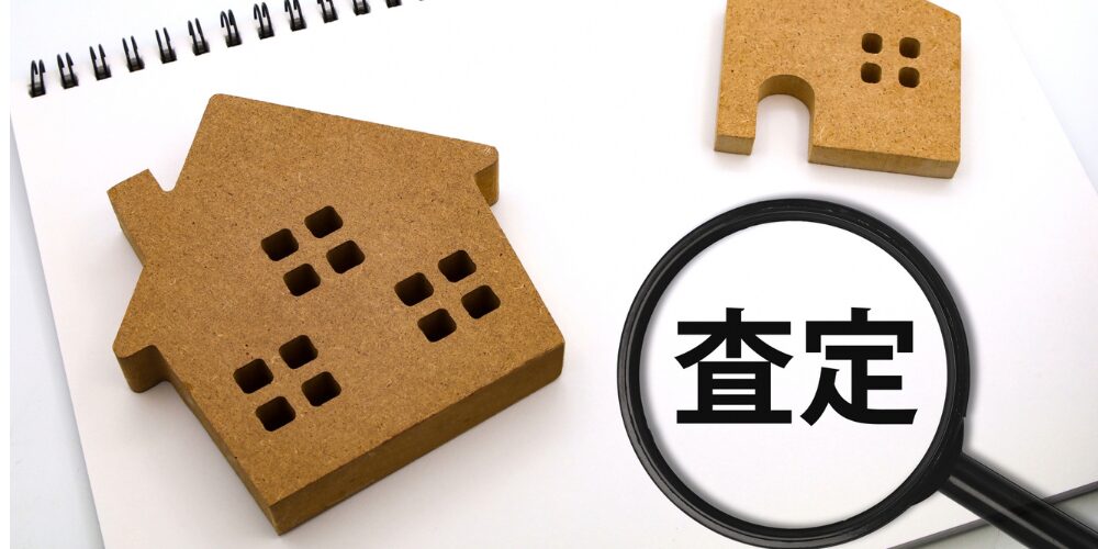スパイラルノートの上に二つの木製家型オブジェと、拡大鏡で黒字の『査定』を拡大表示した構図