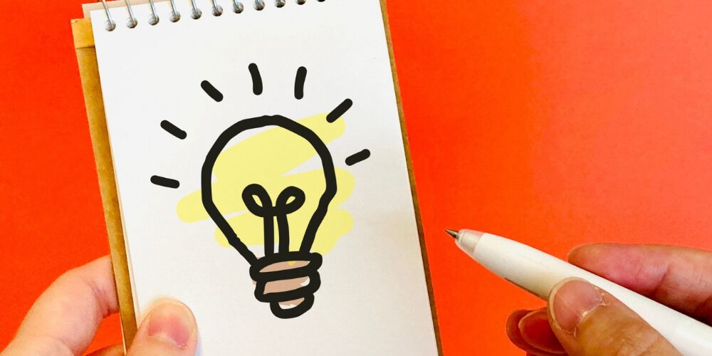 赤い背景の上で手に持った小さなリングノートの白いページに、手描き風の黄色い電球イラストが描かれ、もう一方の手がペンを構えている光景