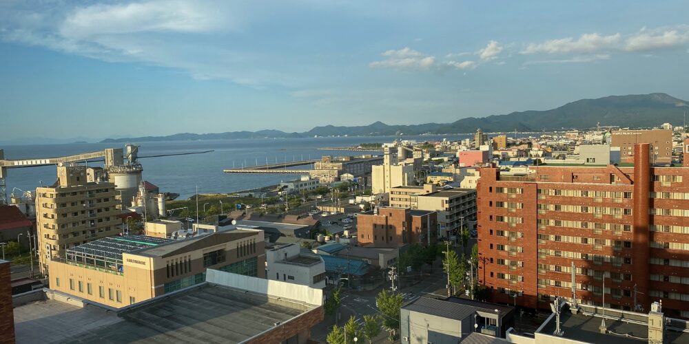 高層ビルやマンションが立ち並ぶ街並みと、その先に広がる静かな海、遠くに連なる山々を俯瞰でとらえた風景