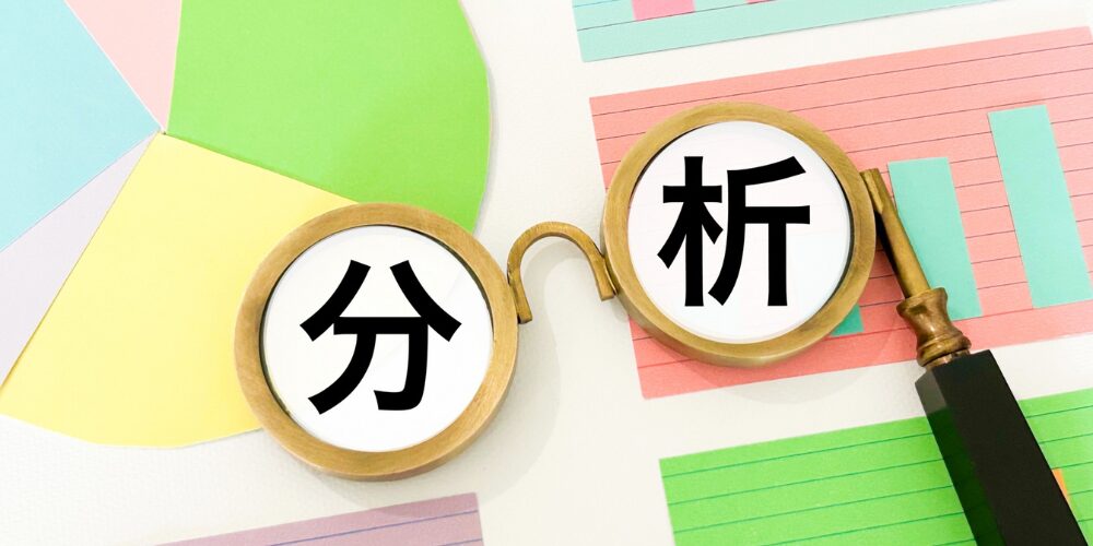 色とりどりの円グラフや棒グラフの用紙の上に置かれた丸い虫眼鏡型フレームに、左側に「分」、右側に「析」と黒字で書かれているビジュアル