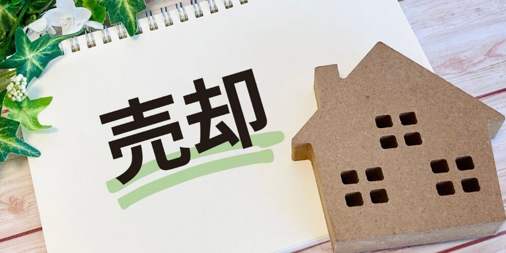 リングノートを開いた白い紙面に緑線で下線を引かれた黒字の『売却』の文字と、その横に木製の家型オブジェが置かれている様子