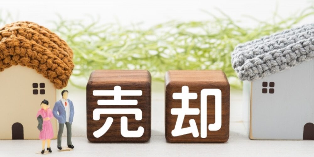 不動産売却を象徴する家の模型と「売却」と書かれた木のブロック、家族のミニチュアが並ぶイメージ写真