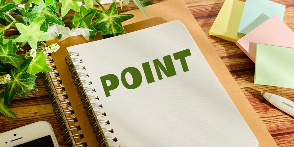 木目のデスク上に置かれた観葉植物と付箋、中央のノートに大きく「POINT」と書かれた構図