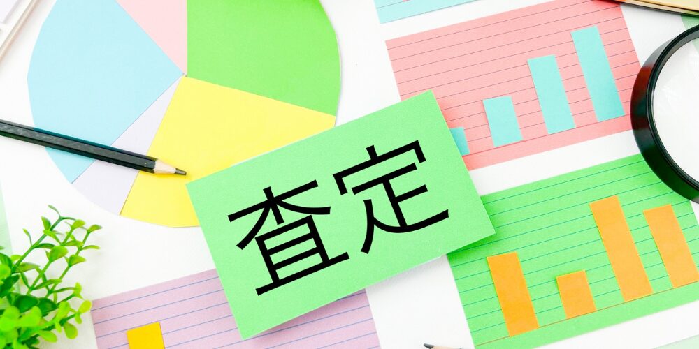 カラフルなグラフ用紙が敷かれた背景に、緑色のカード中央に「査定」と太字で書かれた構図