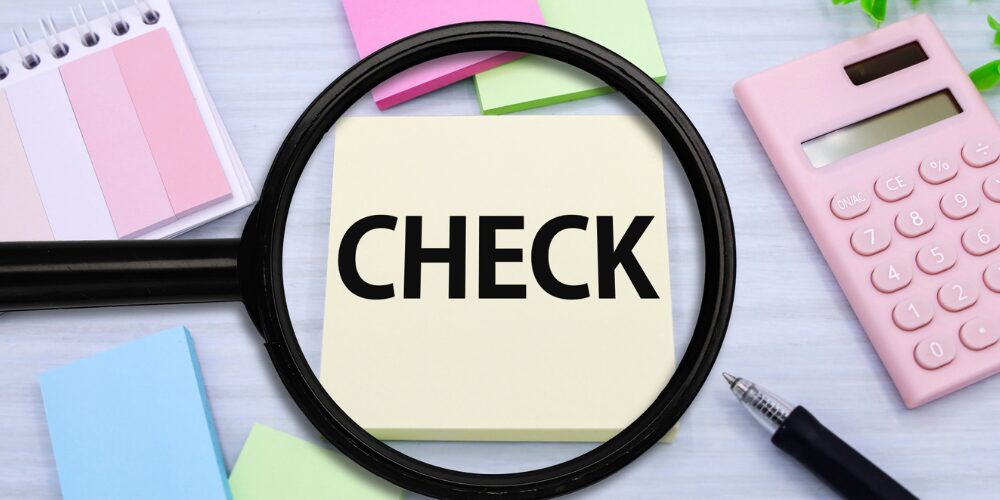 パステルカラーの付箋や電卓、ペンが置かれた机上に、拡大鏡越しに黄色いメモ用紙の『CHECK』の文字がくっきり見える構図