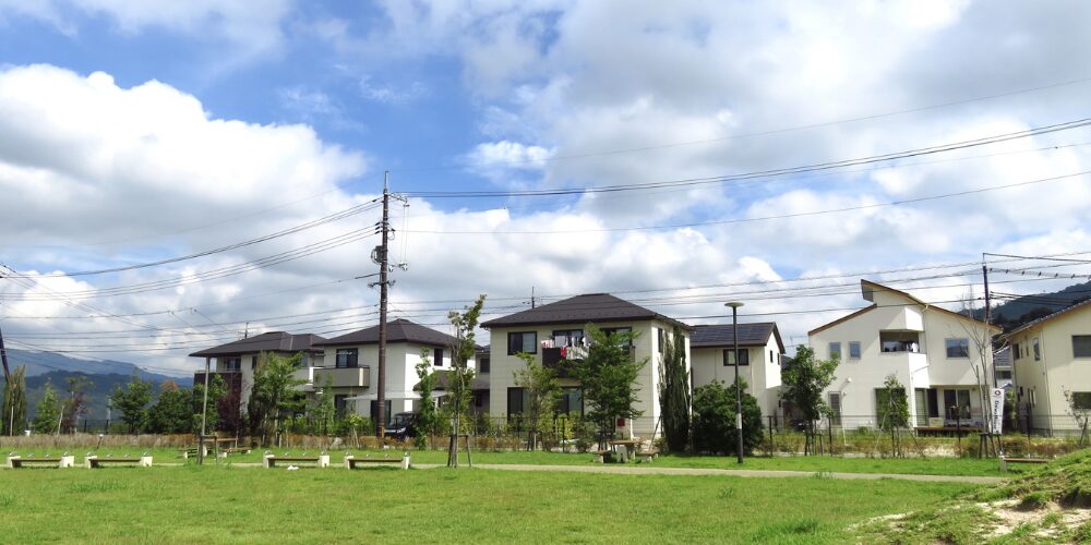 青空の下、広い芝生の手前にベンチが並び、その奥に白やベージュの一戸建て住宅が立ち並ぶ分譲地の風景。