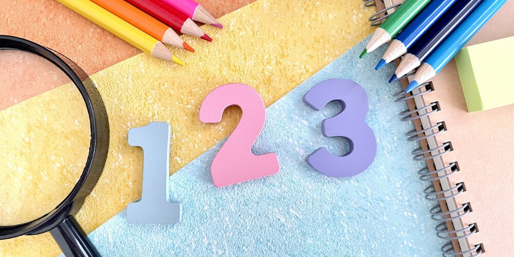カラフルな色鉛筆と虫眼鏡、そしてパステルカラーの『1』『2』『3』の数字がカラフルに並び、横にスパイラルノートが見える文房具の平置き写真