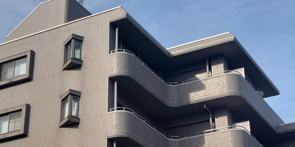 グレーのタイル貼りマンションの角部分を見上げた構図。曲線を描くバルコニーが連続している。