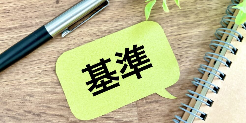 木製のデスクにノートとペンが置かれ、黄緑色の吹き出し型付箋に「基準」という文字が書かれています。