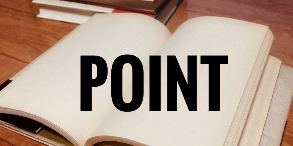 開きかけの書籍の白ページに黒字で大きく『POINT』と書かれている様子。背景には複数の付箋や文房具が並ぶ