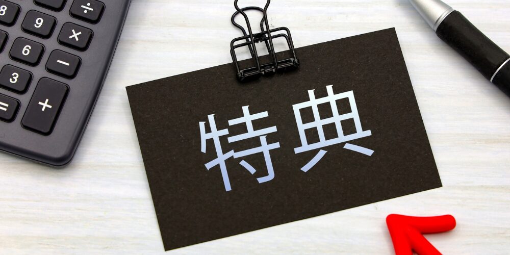 黒いクリップで留められた黒いカードに白字で『特典』と書かれ、隣には電卓と赤い矢印オブジェが置かれたデスク上の光景