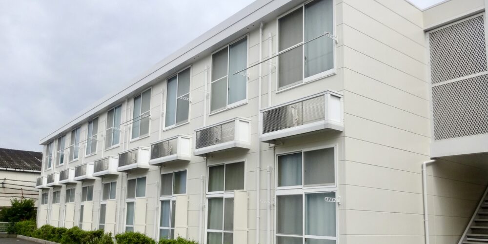 雲の浮かぶ空を背景に、白い外壁とガラス窓が並ぶ二階建てアパートの外観。各部屋にエアコン室外機と物干し金具が設置されている様子