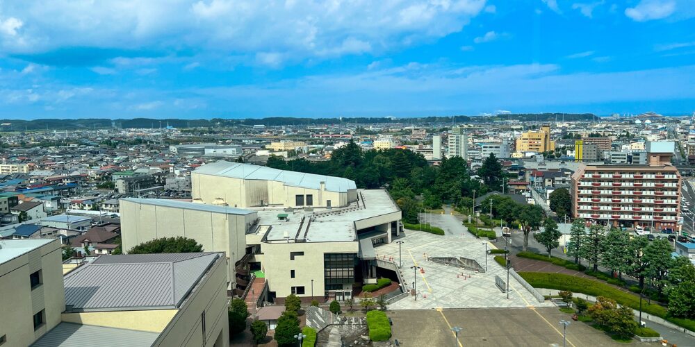 高層ビルの屋上から見下ろす都市のパノラマ。中央手前に大きな公共施設の建物があり、その向こうに住宅街や商業ビルが水平線まで広がる風景。青空と白い雲が空を覆っている様子