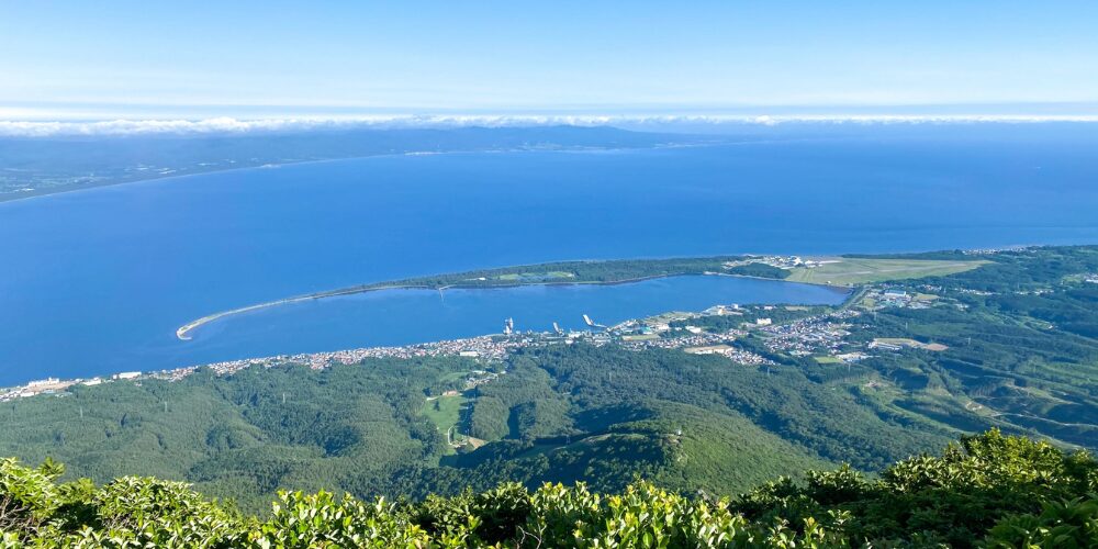 高い場所から見下ろした美しい海岸線の風景。青く広がる海と湾曲した砂浜、森林や街並みが一望できる壮大な景色です。水平線の先には雲がかかった山並みも確認できます。