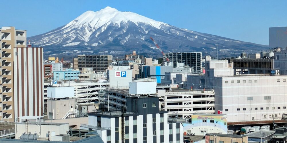 郊外のビル群の先に、雪をいただく大きな山（岩木山）が青空の下にそびえる街並みの風景