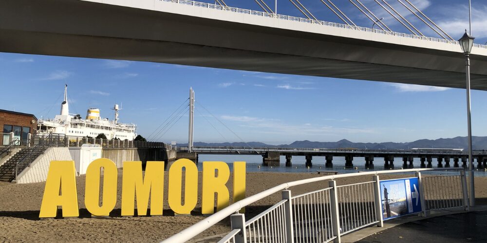 砂浜に設置された黄色い大文字の『AOMORI』オブジェと、その上を跨ぐように架かるシンボリックな橋、遠景に停泊する観光船と建物群
