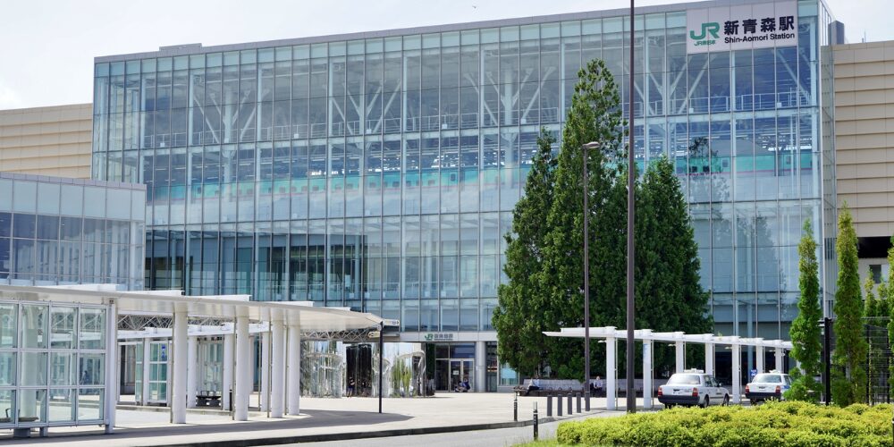 ガラス張りの近代的な駅舎正面。『JR新青森駅 Shin‑Aomori Station』のロゴと文字が見え、手前のタクシーロータリーや緑の植栽が広がる風景