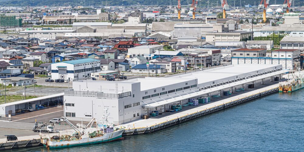 八戸港の岸壁に沿って長く伸びる白い倉庫と荷捌き場、その背後に住宅街や工場の建物群が並ぶ俯瞰の港湾風景