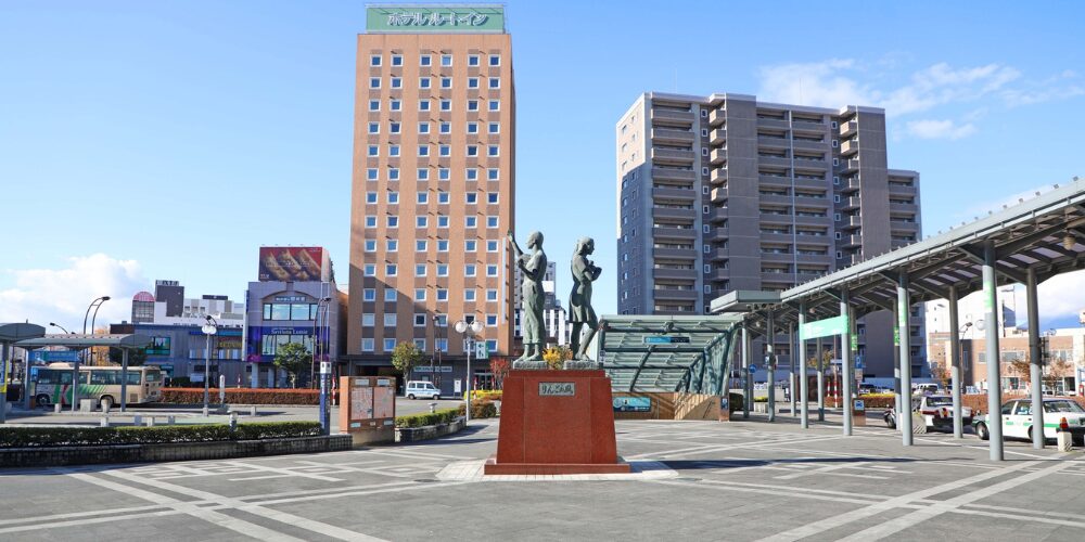 駅前広場のロータリー中央にりんごのモニュメント像、その背後に『ホテルルートイン』の看板を掲げたビルとマンションが建ち並ぶ風景