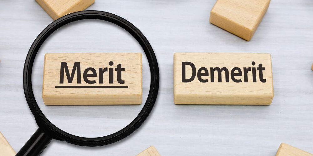 白い背景上に、木製ブロックに「Merit」「Demerit」と書かれ、虫眼鏡が「Merit」の上に重ねられています。