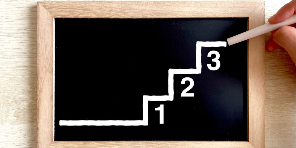 木枠の黒板にチョークで階段状に描かれた線と『1』『2』『3』の数字が書かれ、上部をチョークでなぞる手元が写っている光景