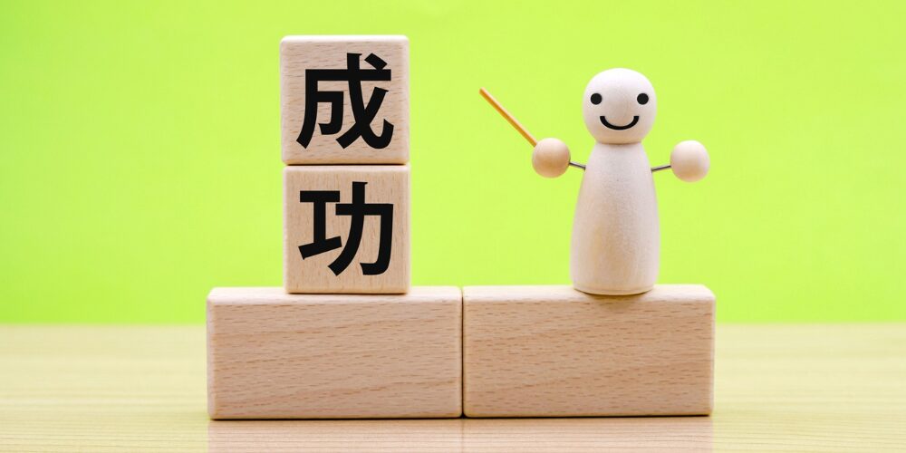 緑色の背景を背に、木製ブロックで「成功」と書かれ、その横で棒を持った木製の人形が微笑んでいます。