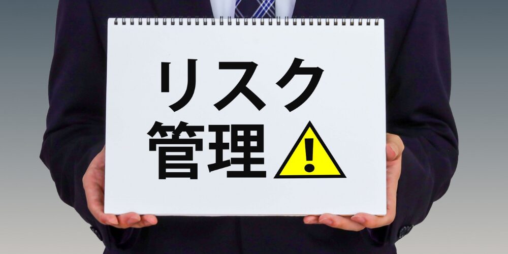スーツを着た人物が「リスク管理!」と書かれたスケッチブックを手に持っています。「!」マークは注意を促す黄色の三角マーク内に記されています。