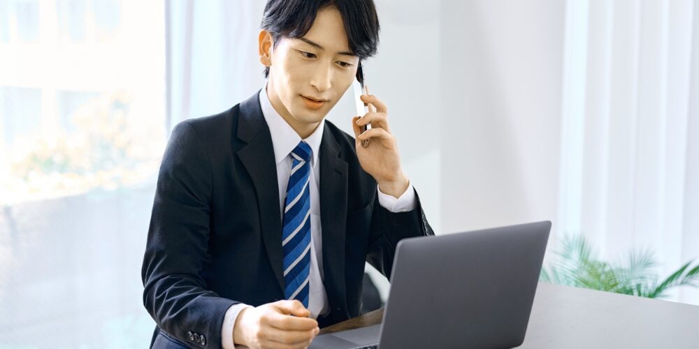 ビジネスマン風の背広姿の男性がオフィス机でノートパソコンを前に、片手でスマートフォンに通話しながら話を聞いている様子