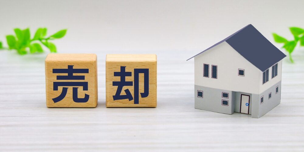 白い木目調テーブル上に、木製ブロックに紺色で『売』『却』と書かれ、横に小さな住宅模型が置かれたイメージ