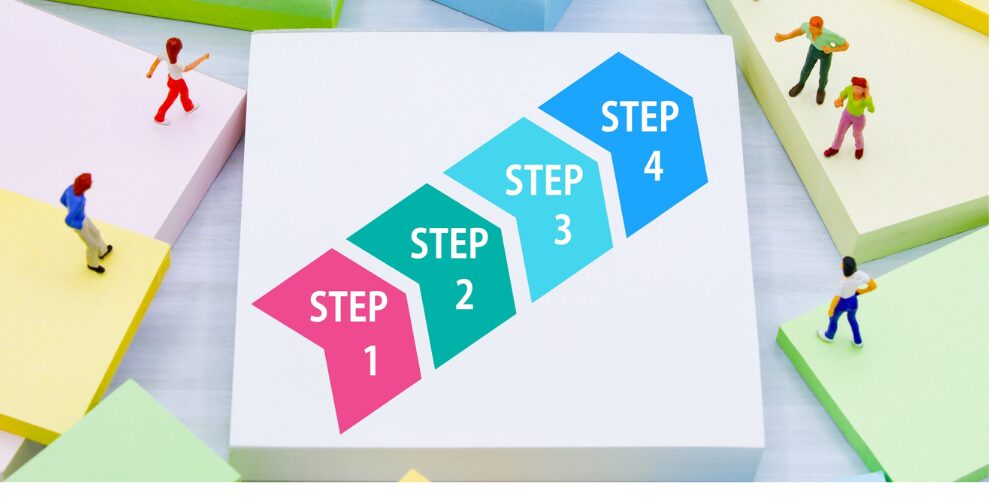 カラフルな台形ブロックが階段状に『STEP 1』『STEP 2』『STEP 3』『STEP 4』と並び、周囲の台座上に小さな人形が歩いているビジュアル